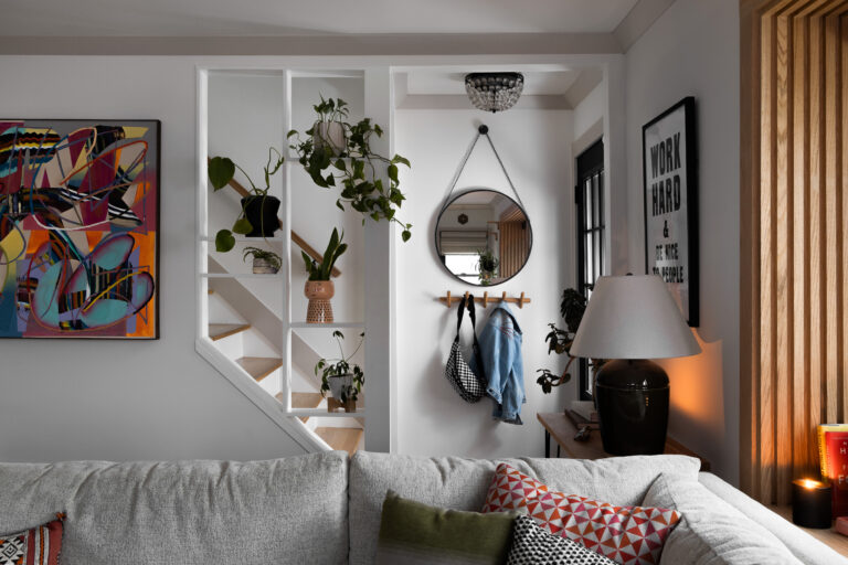 A cozy and eclectic living area featuring an unpainted oak wood trim that adds natural warmth to the space. The room includes a gray couch adorned with colorful throw pillows, a side table with a ceramic lamp, and a framed motivational print on the wall. A small entry nook with a round mirror and hooks for coats creates a functional and stylish entryway. Potted plants add greenery, while an open partition with oak trim provides a subtle division between the living room and staircase, blending modern decor with the classic charm of natural wood accents.