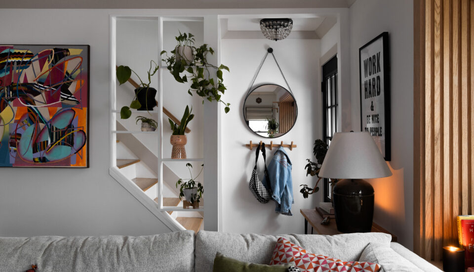A cozy and eclectic living area featuring an unpainted oak wood trim that adds natural warmth to the space. The room includes a gray couch adorned with colorful throw pillows, a side table with a ceramic lamp, and a framed motivational print on the wall. A small entry nook with a round mirror and hooks for coats creates a functional and stylish entryway. Potted plants add greenery, while an open partition with oak trim provides a subtle division between the living room and staircase, blending modern decor with the classic charm of natural wood accents.
