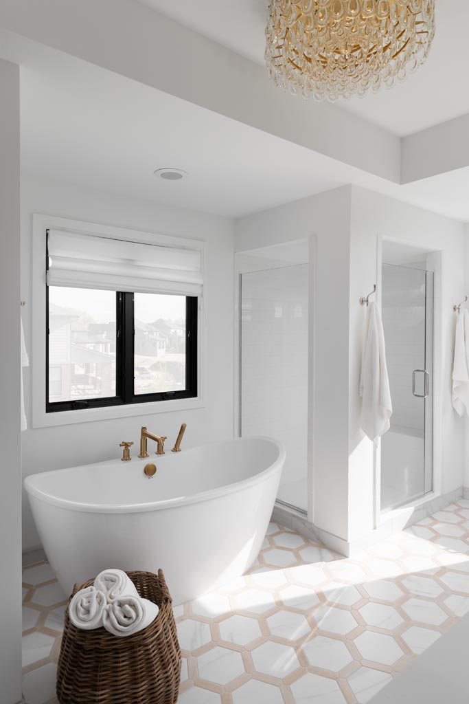 A bright and airy bathroom in Birmingham, Michigan, featuring a freestanding white tub with brass fixtures, geometric floor tiles, a walk-in shower, and a woven basket filled with towels.