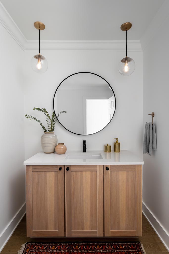 Minimalist spa-like powder room with a white oak vanity, round mirror, pendant lighting, and brass accents.