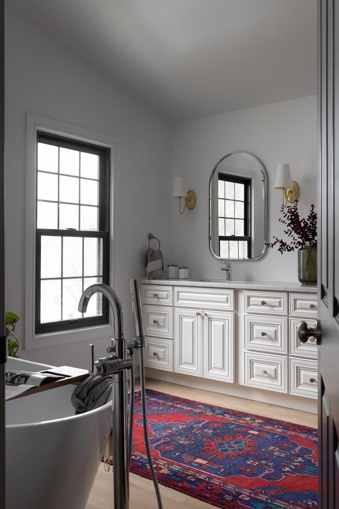 Bathroom renovation featuring a freestanding bathtub, custom white double vanity, gold accents, and a red rug.