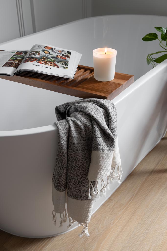 Close-up of a white soaking tub with bath tray, grey towel, and wood flooring in a remodeled Canton, MI bathroom.
