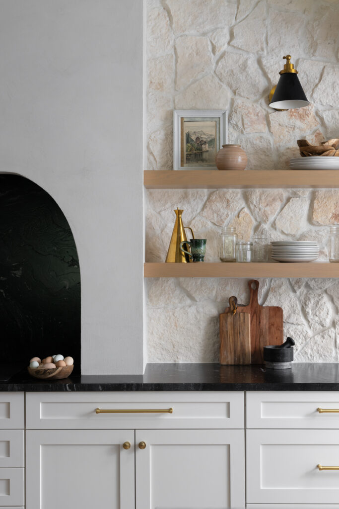  Biophilic kitchen design featuring a natural stone wall, wood open shelving, brass accents, and nature-inspired decor. The space incorporates natural materials to create a warm, inviting environment that promotes well-being and connection with nature.