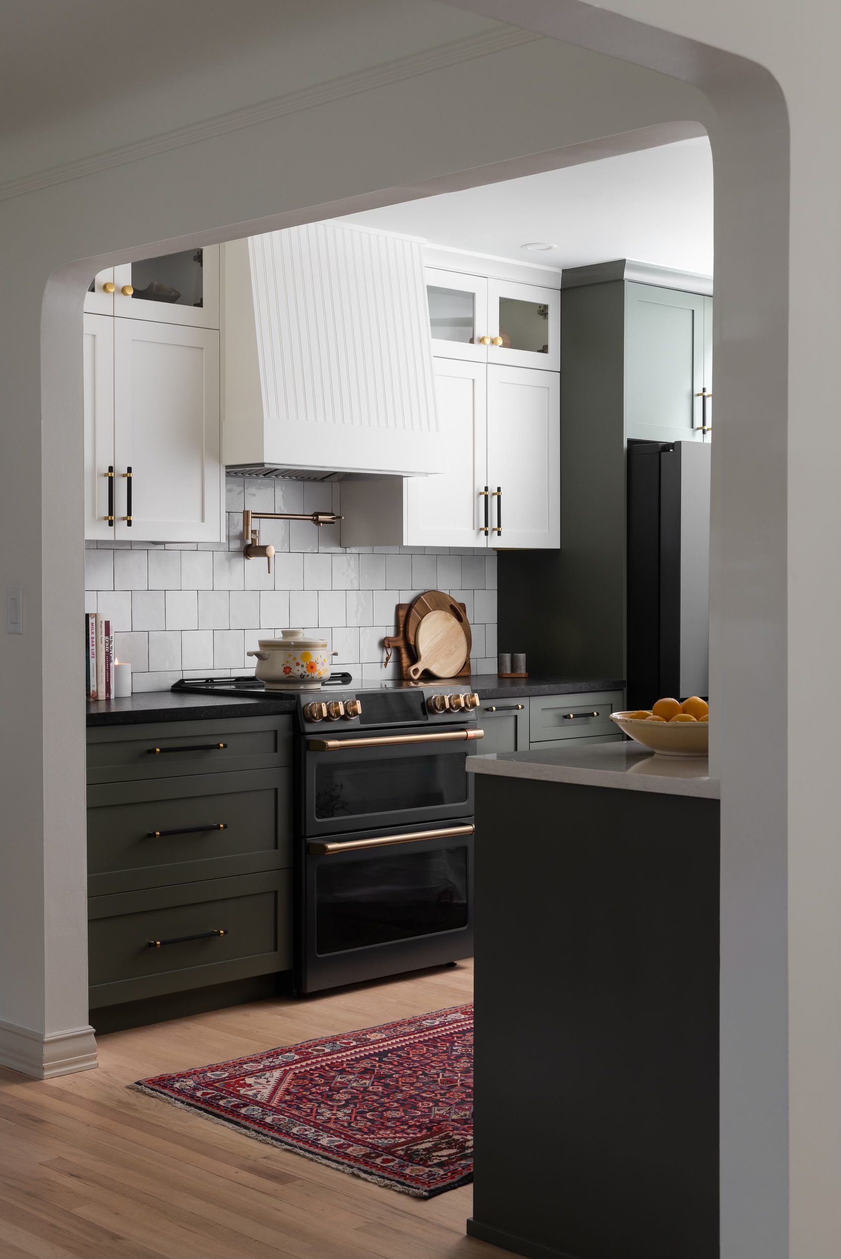  A stylish modern kitchen featuring bold green lower cabinets, white upper cabinets, and brass hardware. The kitchen includes a sleek black range with brass accents, a white tile backsplash, and a colorful rug, adding warmth and character to the space. Perfect for homeowners looking to incorporate bold color palettes and luxury materials into their kitchen design.