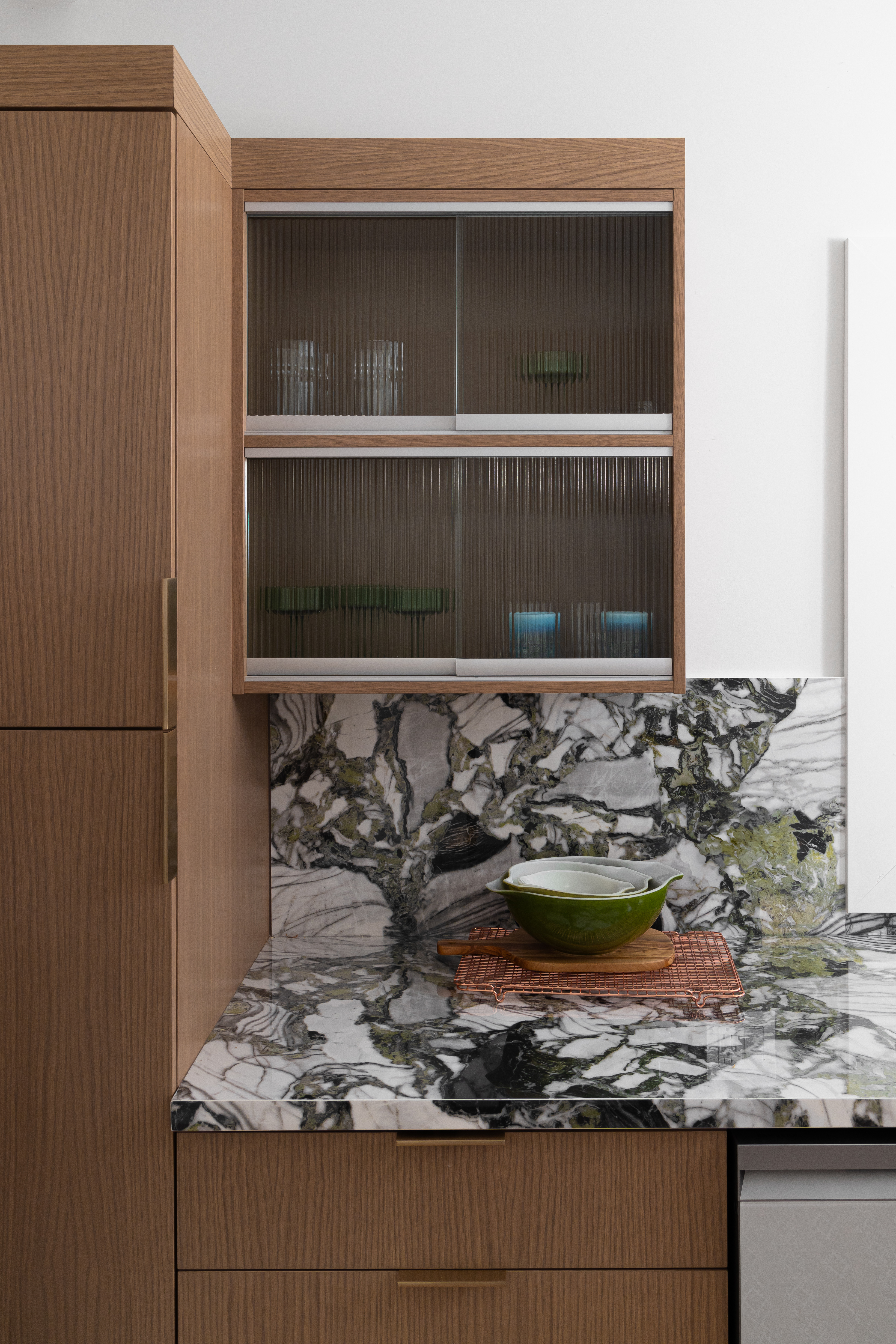 Natural wood cabinetry paired with fluted glass shelving and bold marble countertops featuring green accents in a luxury kitchen design.