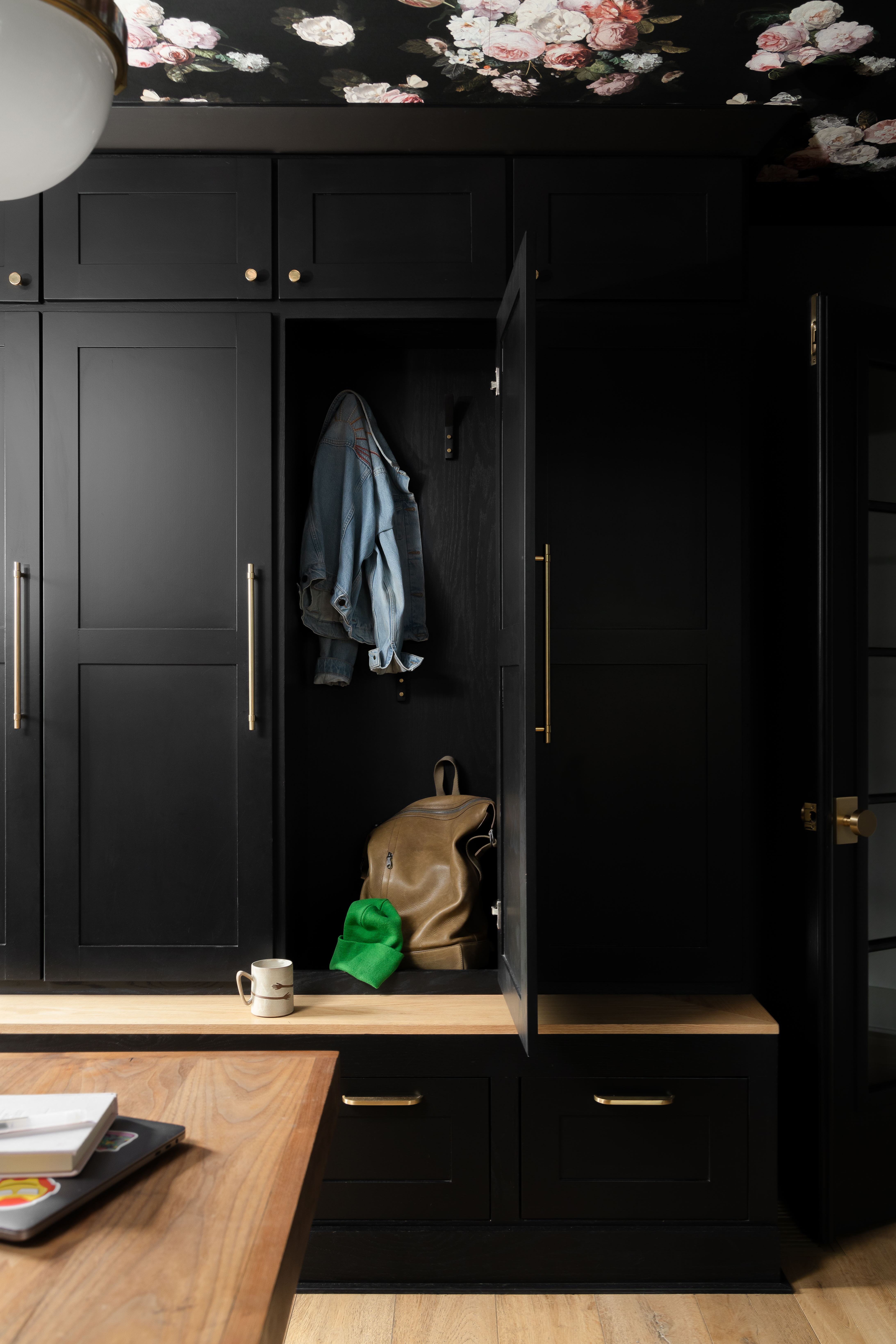 Modern mudroom with black cabinetry, brass hardware, and a floral ceiling. Features a built-in wooden bench and storage with open locker space for hanging items, creating a stylish and functional entryway.
