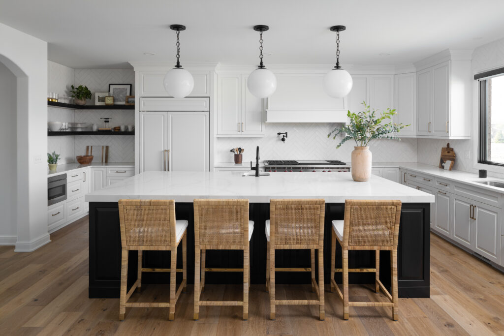 A modern luxury kitchen featuring a large island with a black base, white marble countertop, and woven bar stools. Three large, globe pendant lights provide statement lighting over the island. The kitchen has white cabinetry, a built-in refrigerator, and a herringbone tile backsplash, creating a sophisticated and stylish space ideal for homes in Ann Arbor, Michigan.