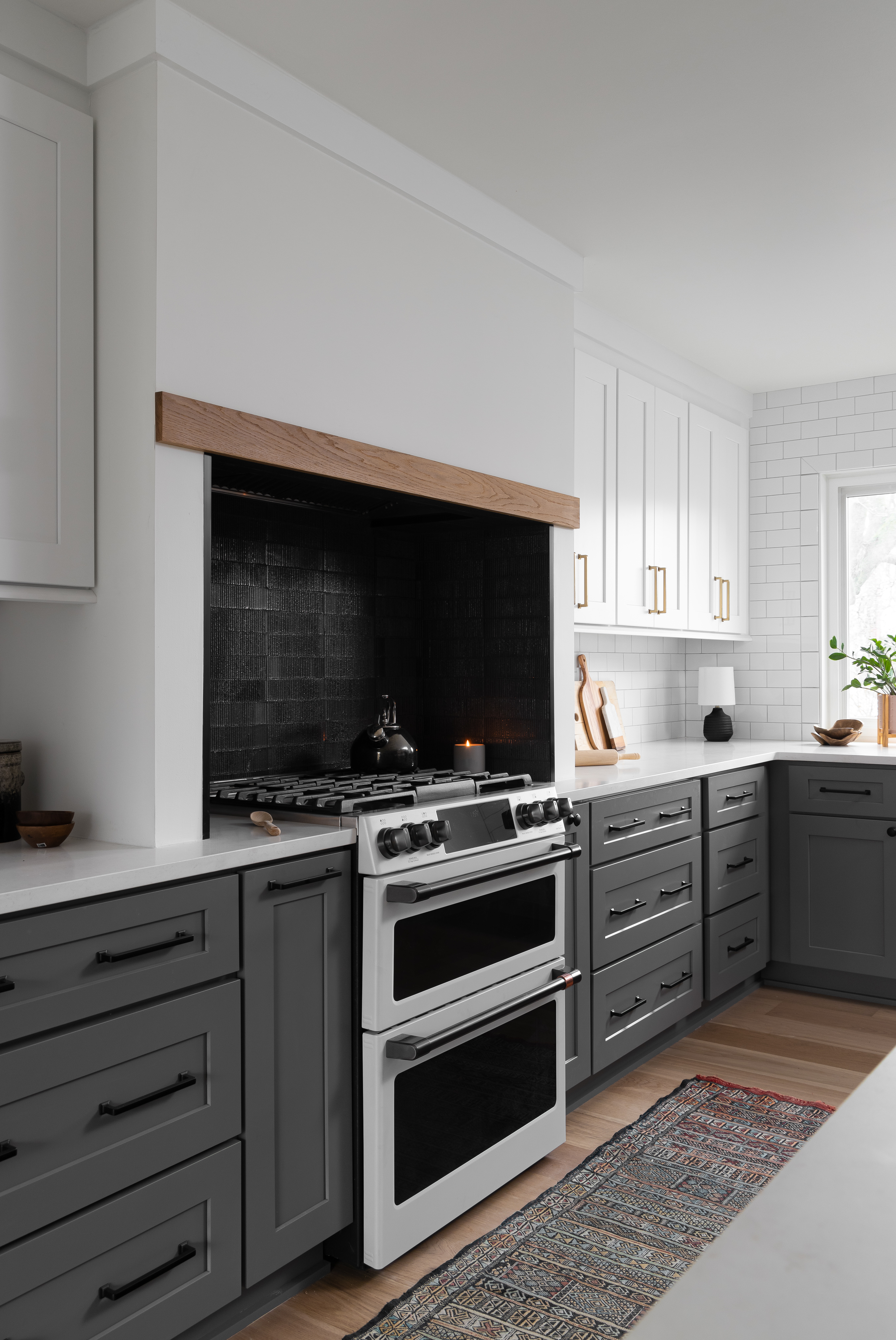 A modern luxury kitchen featuring dark gray lower cabinetry, a black textured backsplash, and a white upper cabinet setup, providing a striking contrast. The kitchen is equipped with a high-end gas range, wood accent details, and an elegant area rug for added warmth. This Southeast Michigan kitchen, designed by Clark + Aldine, combines functionality and style, ideal for homeowners in Ann Arbor and Plymouth.