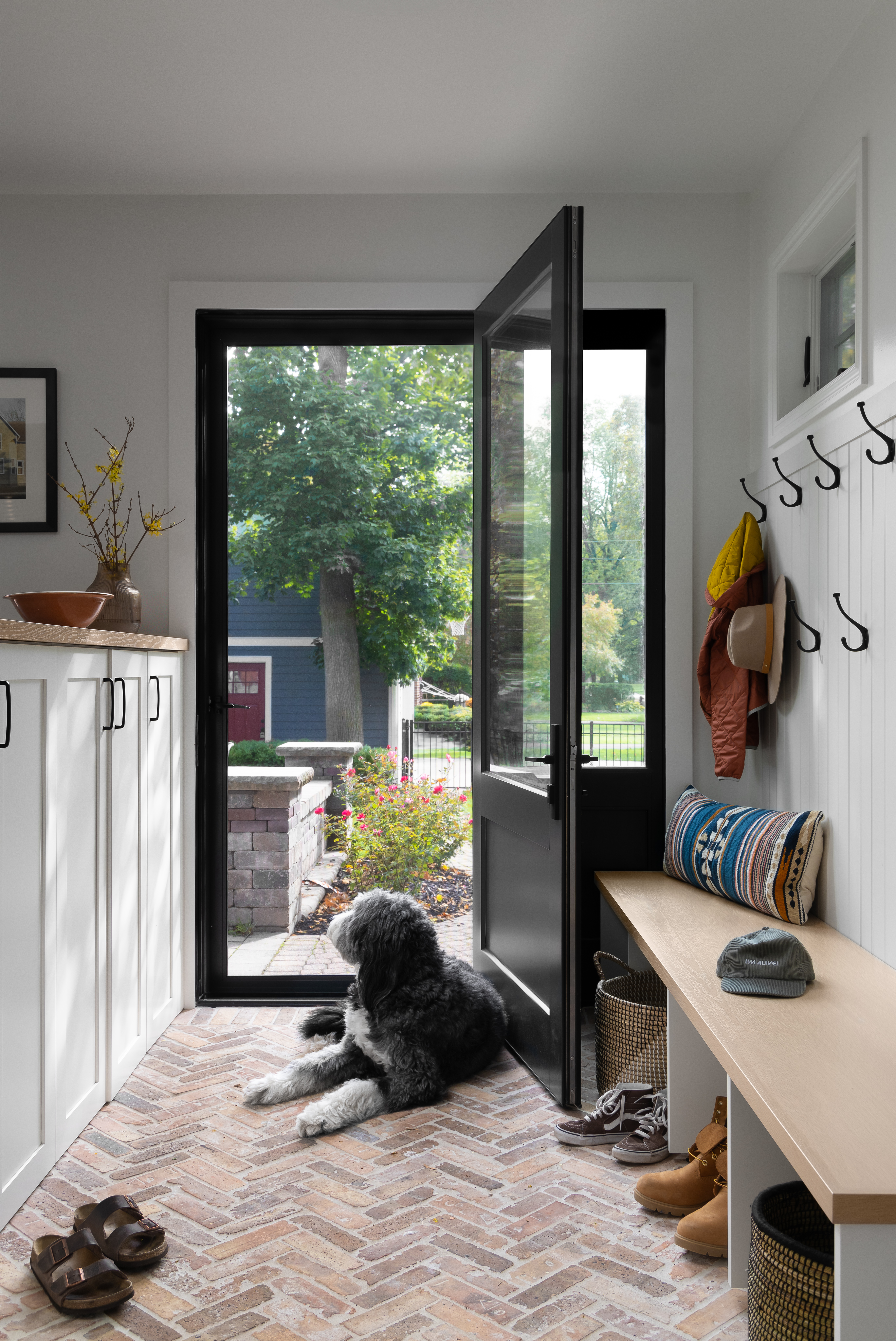 Bright mudroom with herringbone brick flooring, a built-in wooden bench, hooks for jackets and hats, and ample storage space, featuring a dog resting near the open door, creating a welcoming and pet-friendly entryway.