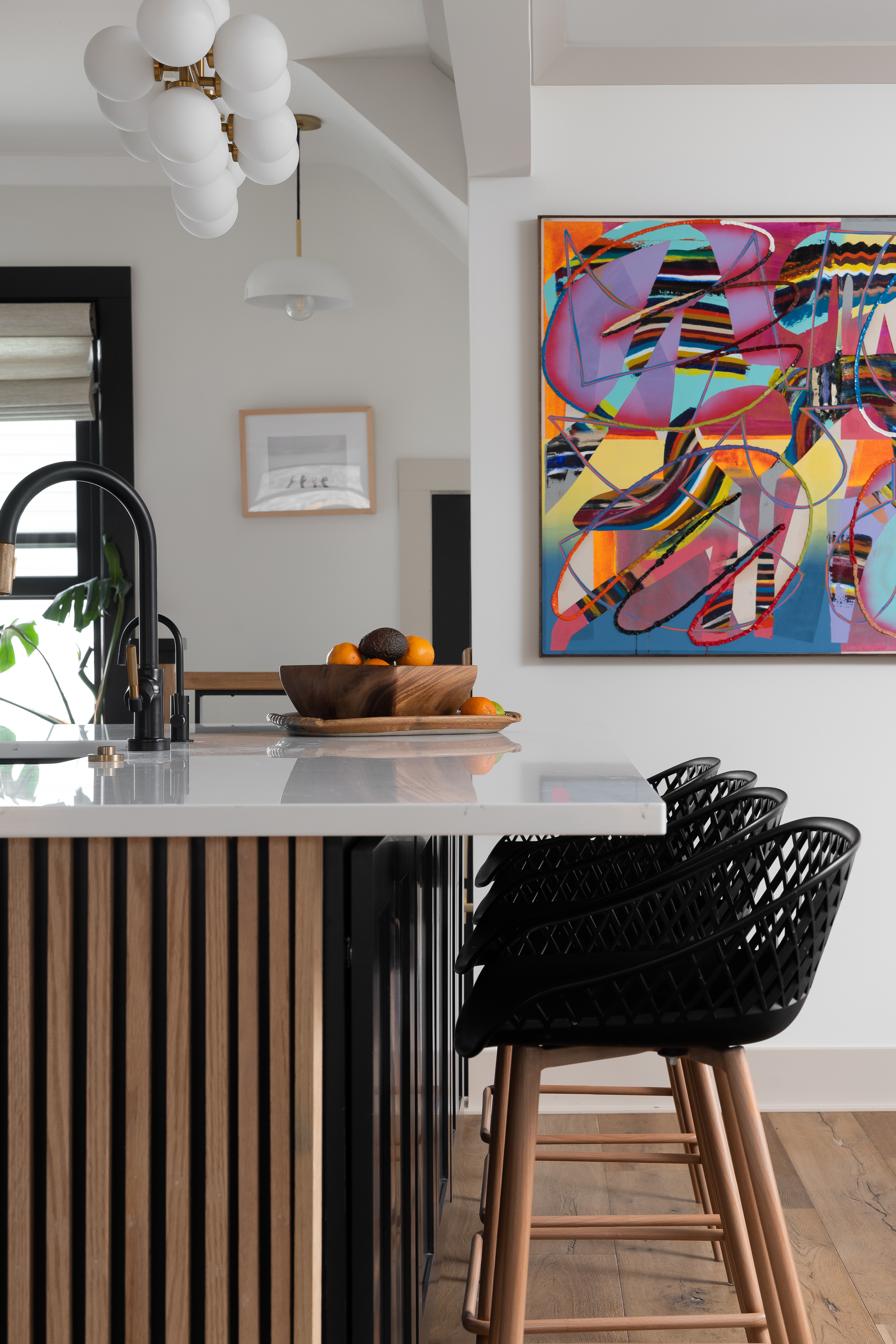 A stylish modern kitchen featuring integrated technology with a sleek black faucet, a white quartz countertop, and wooden slat accents on the island. Black mesh bar stools and a vibrant abstract painting add a pop of personality, blending functionality with contemporary art for a unique kitchen design. Ideal for Ann Arbor, Michigan homeowners seeking a fusion of modern technology and artistic expression.