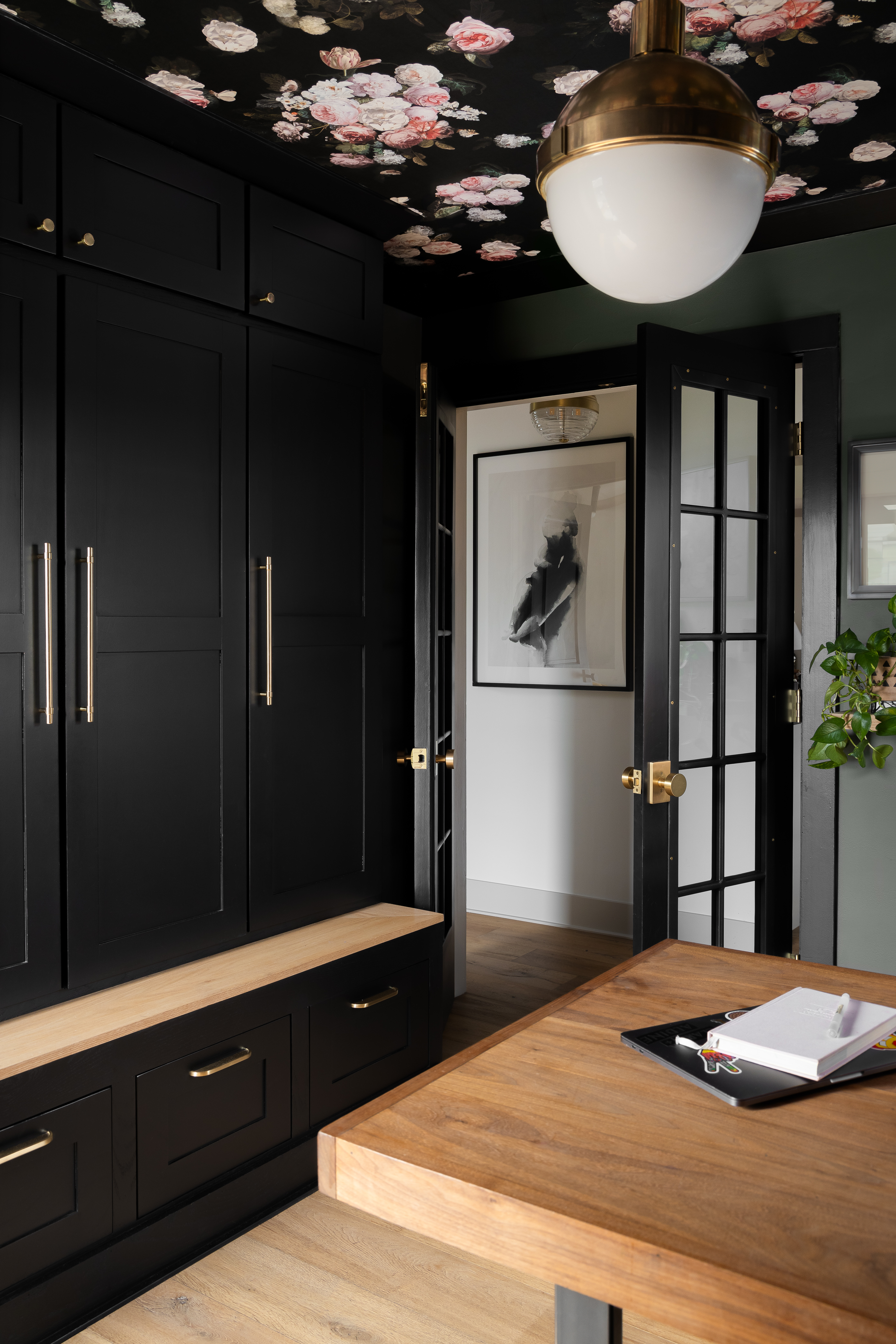 Sophisticated mudroom design featuring black cabinetry with brass hardware, a built-in wooden bench, floral ceiling wallpaper, and French doors for a stylish and functional entryway.