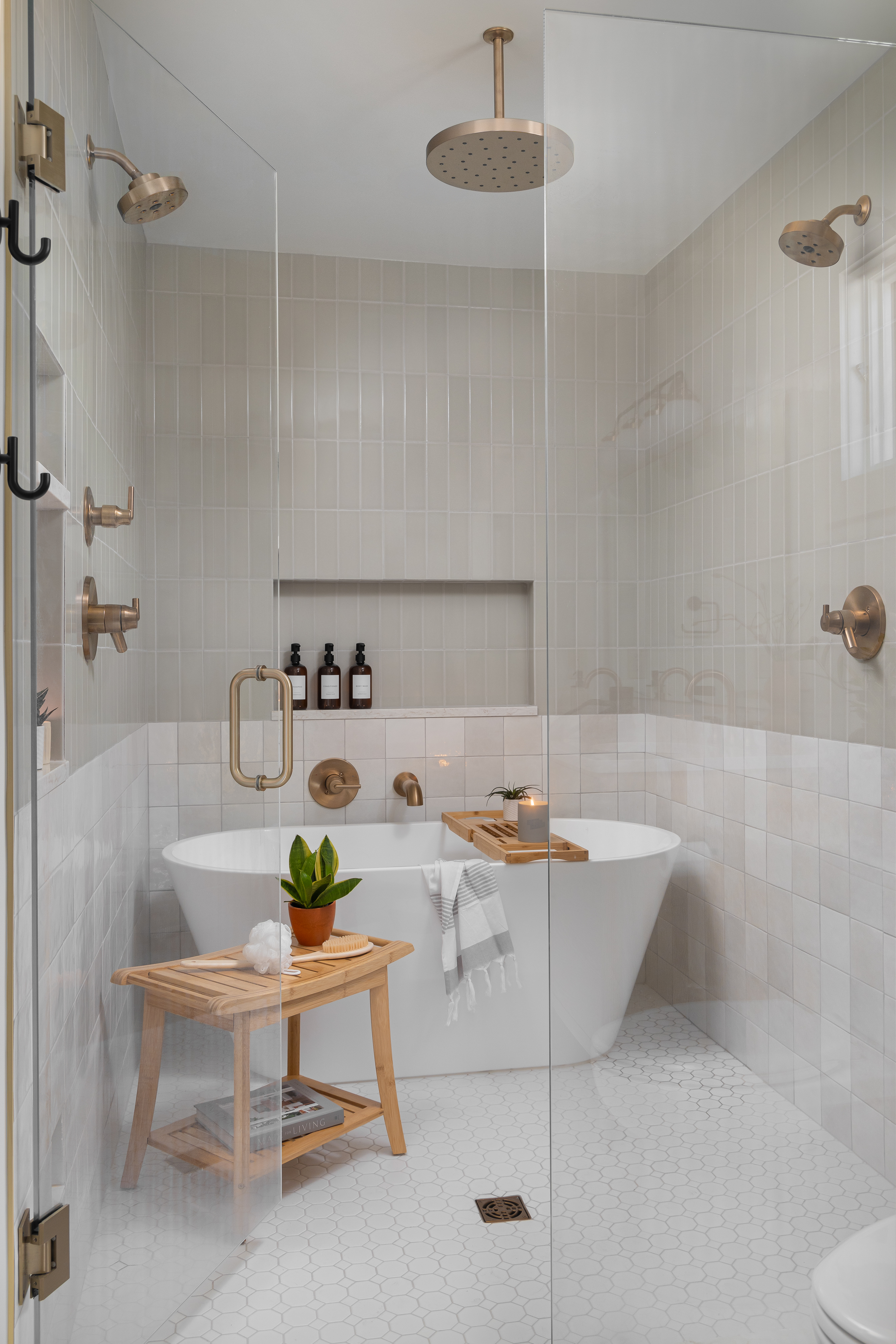 A luxurious spa-like bathroom with a freestanding white tub, brass fixtures, soft beige tile, and a wooden stool topped with greenery and spa essentials in a Michigan lake house.